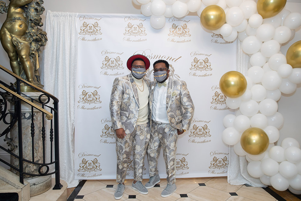 Shonathan and Desmond pose for photos in front of the custom designed logo wall and balloon installation at their engagement party.