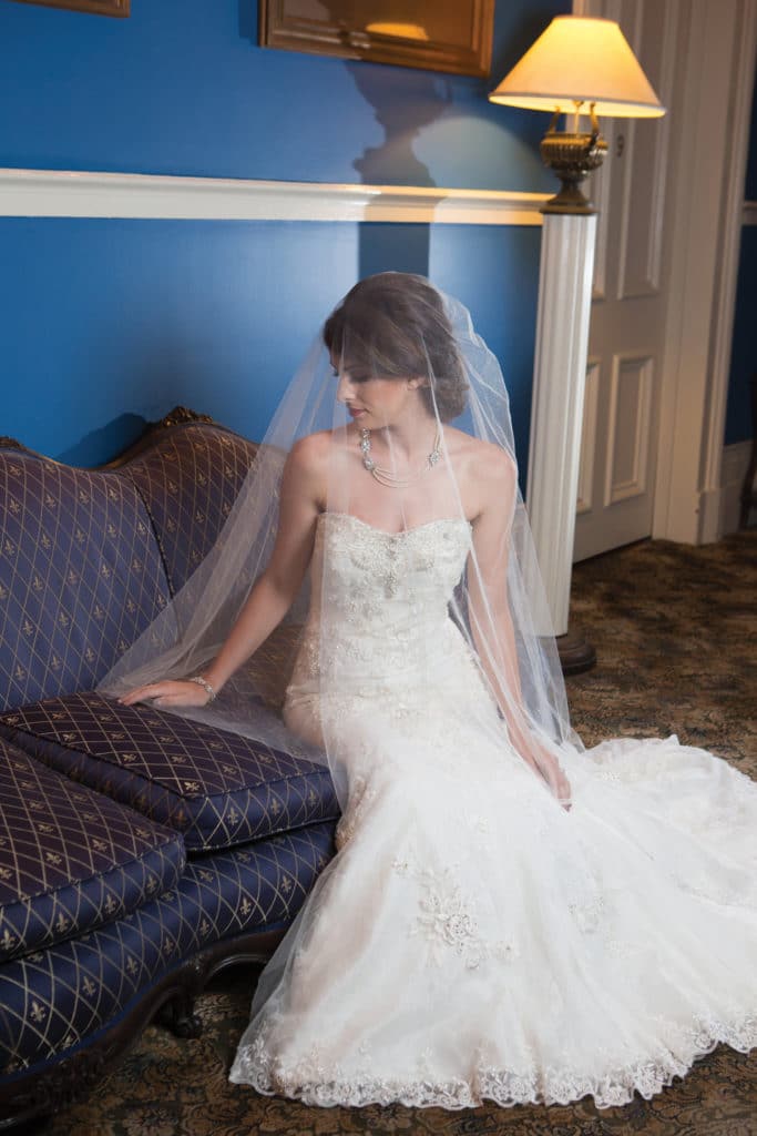 Bride in strapless white gown and veil sits on blue patterned sofa, looking down with hand on cushion in blue-walled room.