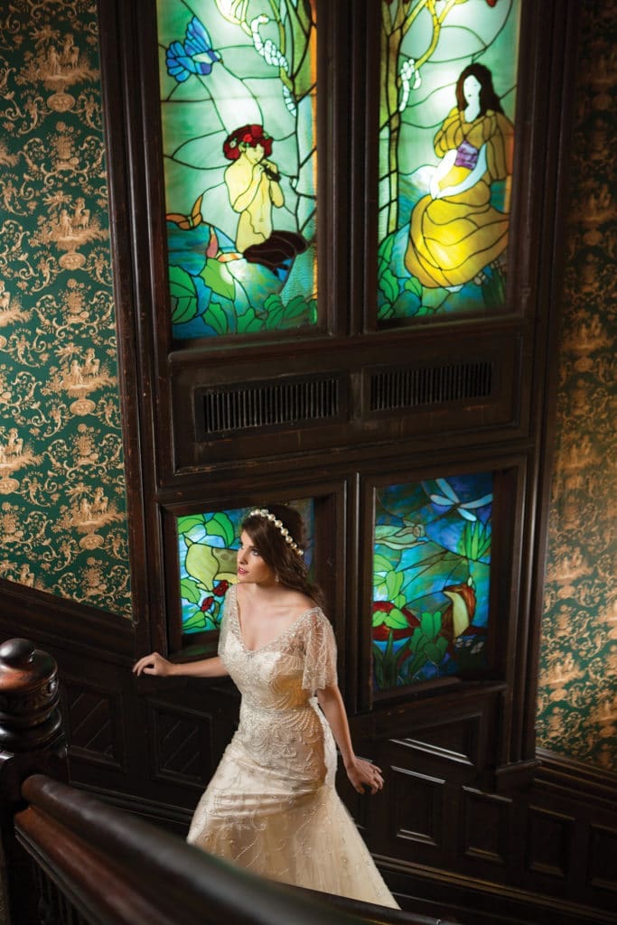 Bride in lace gown ascends wooden staircase, vibrant stained glass of women and flowers in the background.