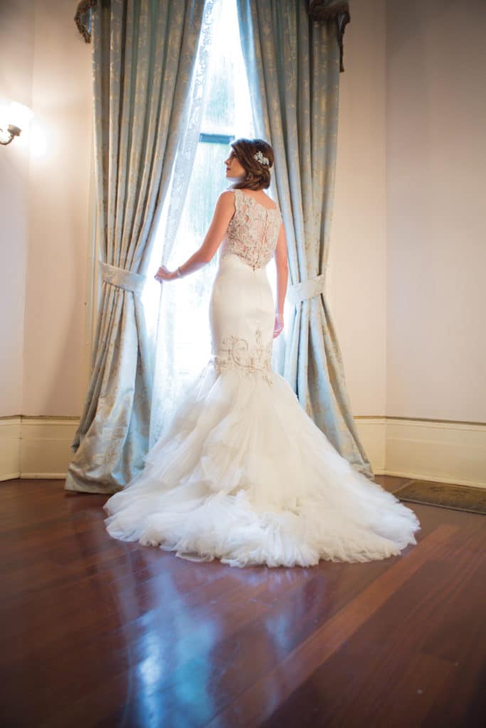 Bride in a white lace-back gown stands inside, holding blue patterned curtains open, gazing out the window.