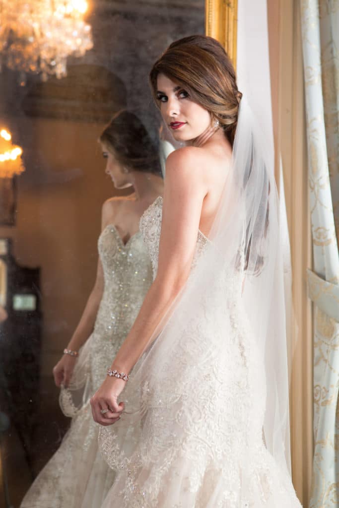 Bride in strapless lace gown with long veil stands by mirror, soft lighting and chandelier reflections behind her.