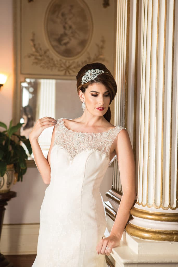 Bride in white lace wedding dress with jeweled headpiece by ornate white and gold columns in a softly lit classic room.