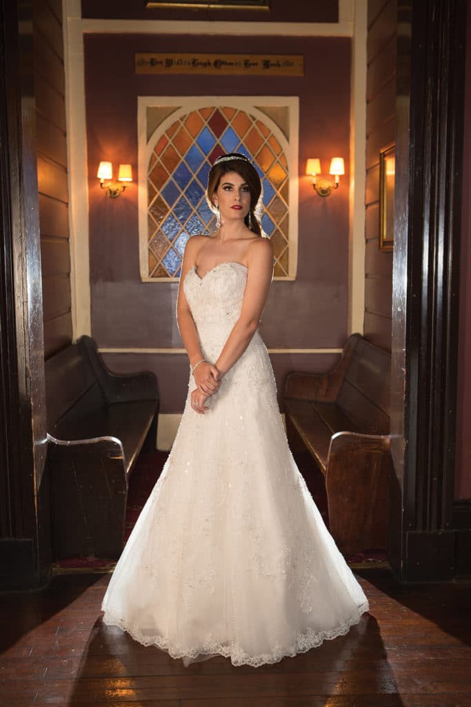 Bride in a strapless white wedding dress standing in a dim room with pews, stained glass, and wall sconces behind her.