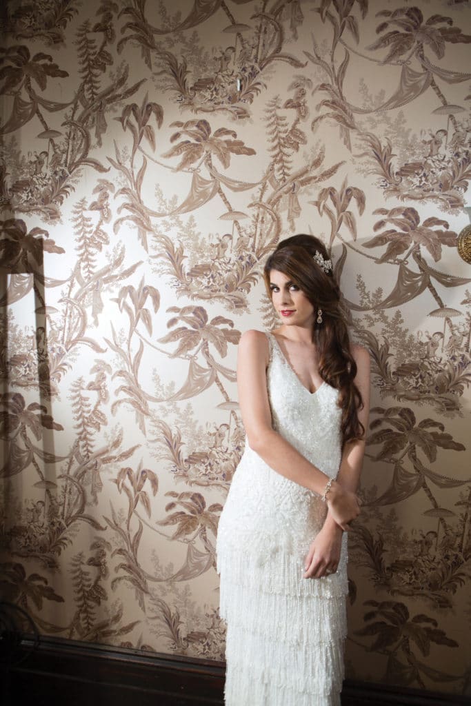 Woman in a white sleeveless dress stands by beige floral wallpaper, looking sideways with hands gently clasped.