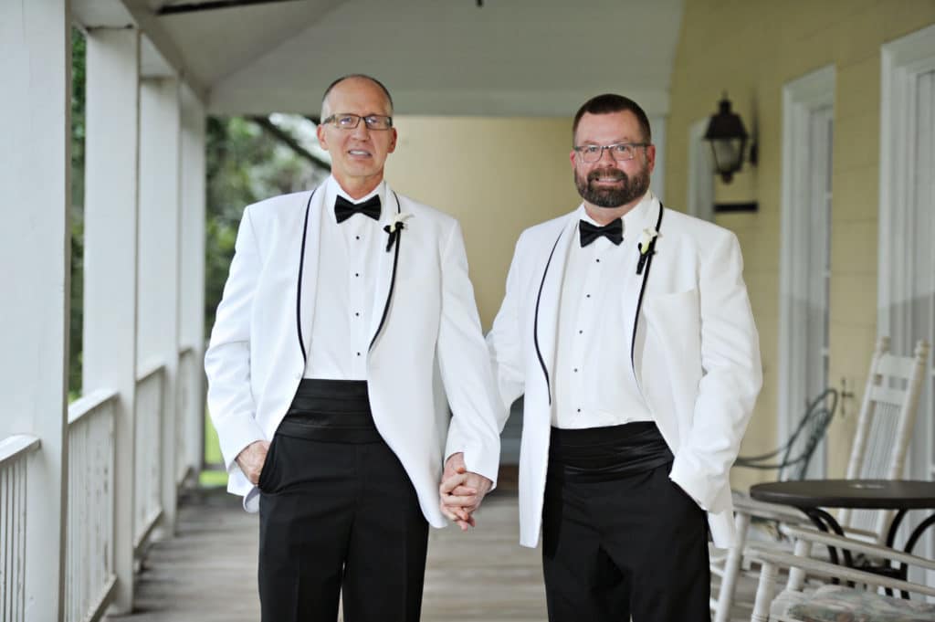 Two men in matching white tuxedos hold hands and smile on a porch with white rocking chairs and a yellow wall behind them.