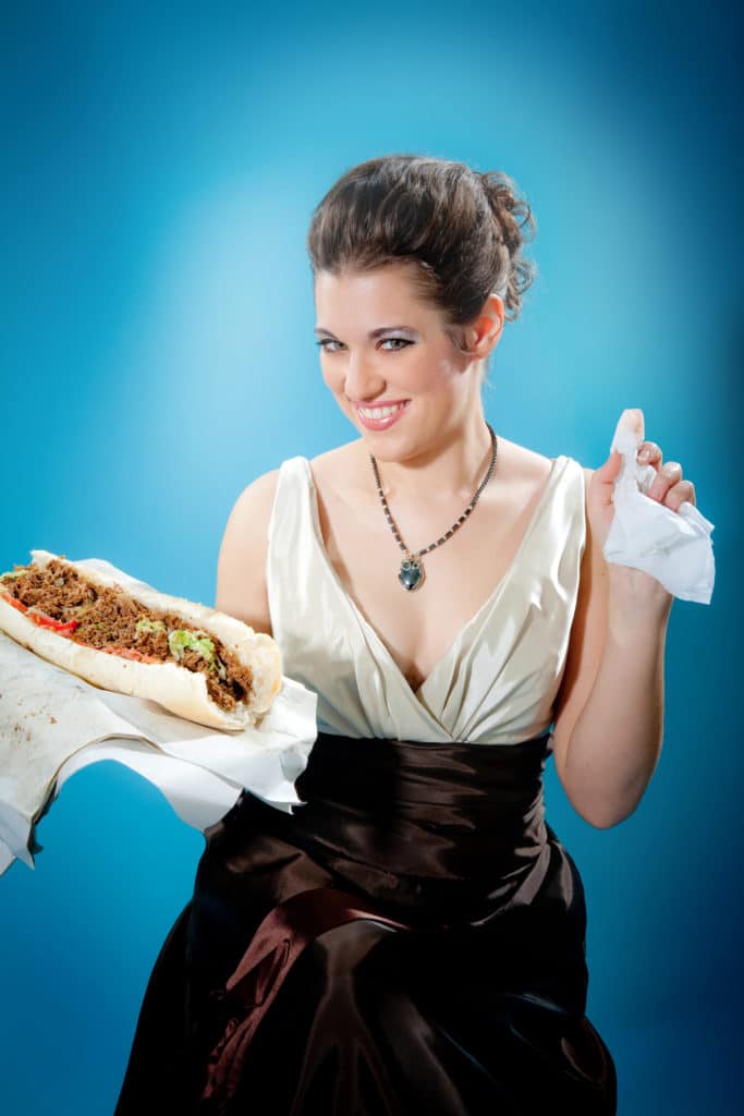 Smiling woman in formal dress holds a large sandwich and napkin, seated against a blue background.