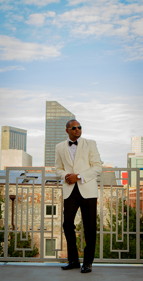Jarrel is wearing a classic shawl lapel ivory dinner jacket ensemble with a Jack Mason Nautical Chronograph watch, Randolph Aviator Sunglasses and gold crown cufflinks from John’s Tuxedos and a gray and rose gold band from Brilliance in Diamonds.