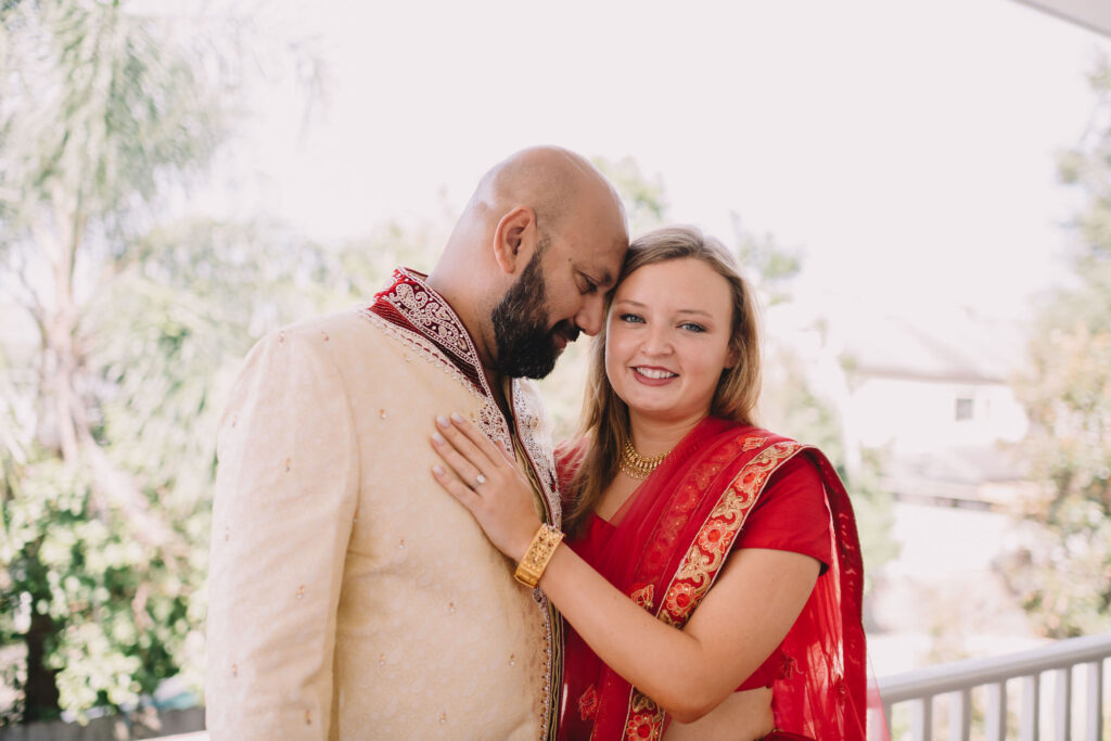 Smiling couple in Indian attire on a sunny balcony, woman in red saree, man in cream sherwani; trees and houses behind.