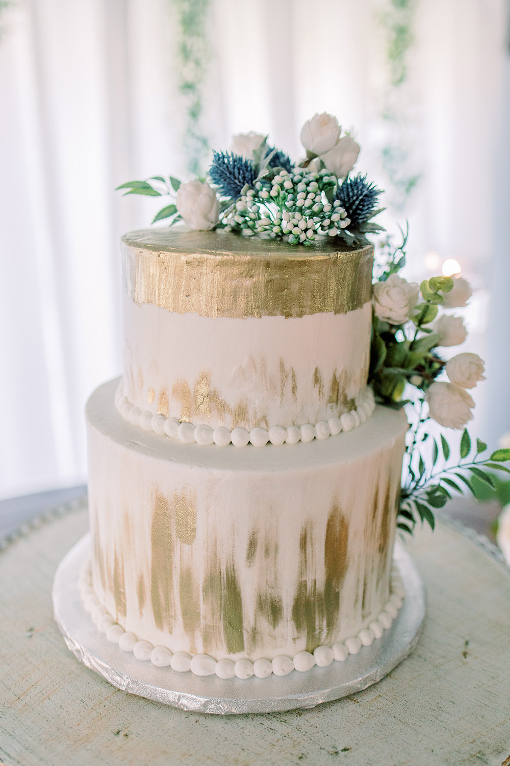 Detail of the Wedding Cake. A two-tier ivory cake with gold accents and fresh flowers and greenery. Photo: Ashley Kristen Photography