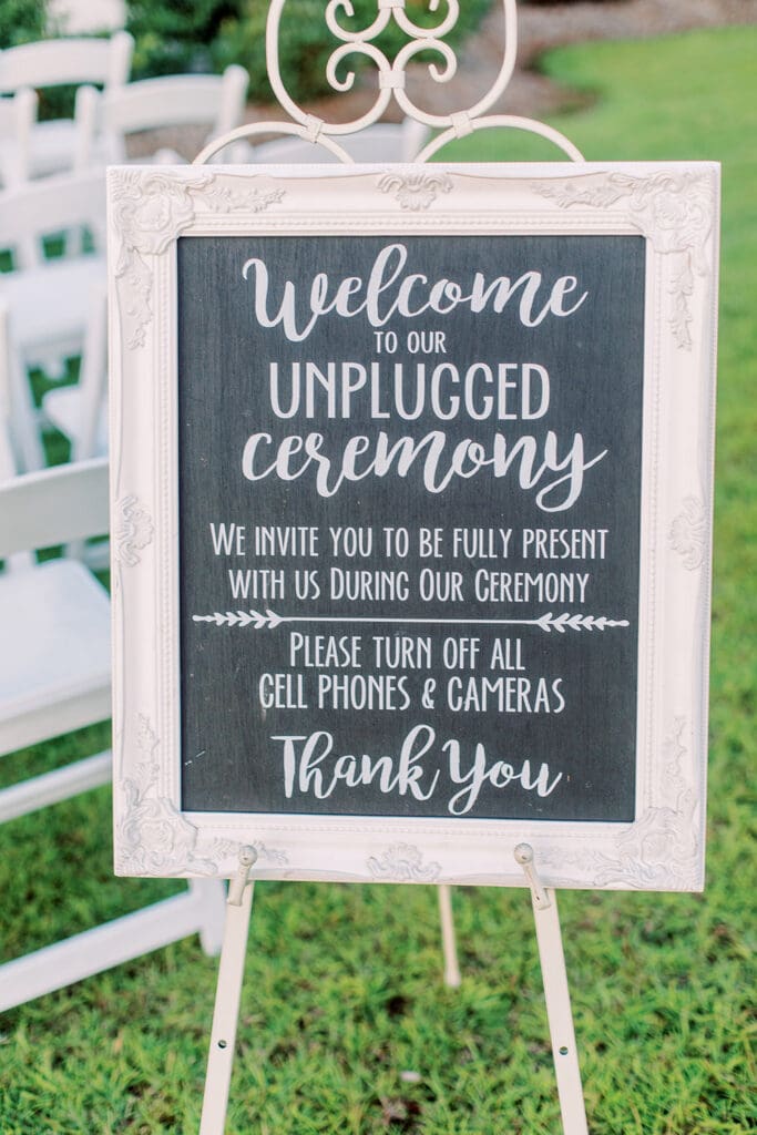 Outdoor Louisiana wedding sign reads “Welcome to our unplugged ceremony.” White chairs and grass set the heartfelt scene.