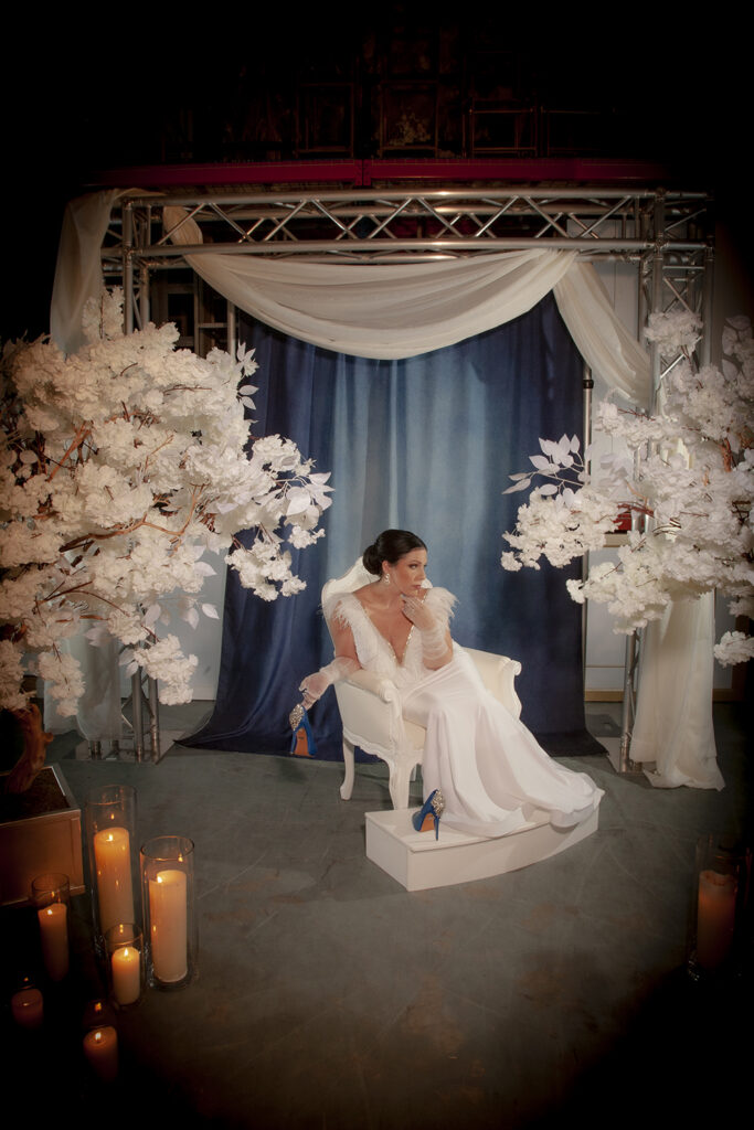 Woman in a long white dress sits on a white chair, surrounded by white flower trees, candles, and draped blue and white fabric.