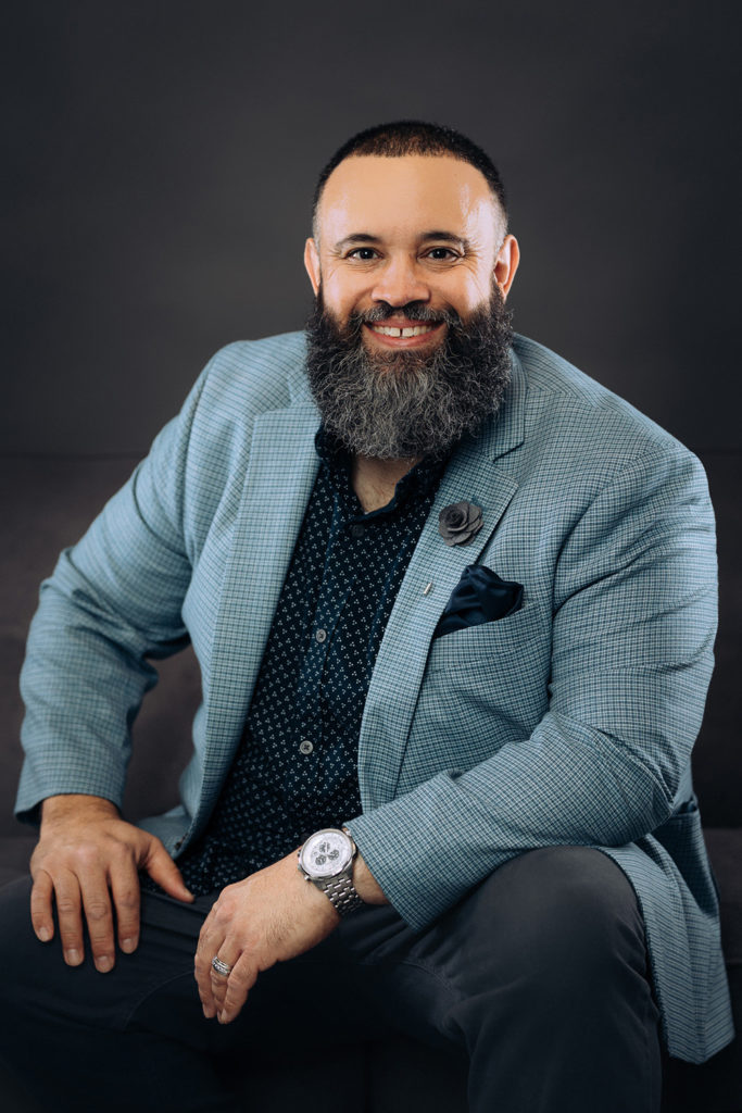 Smiling bearded man in a light blue blazer, dark shirt, and watch sits against a dark background.