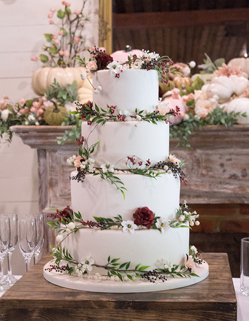 Four-tier white wedding cake with red and white flowers, green leaves, on a wooden stand; glasses and florals in background.
