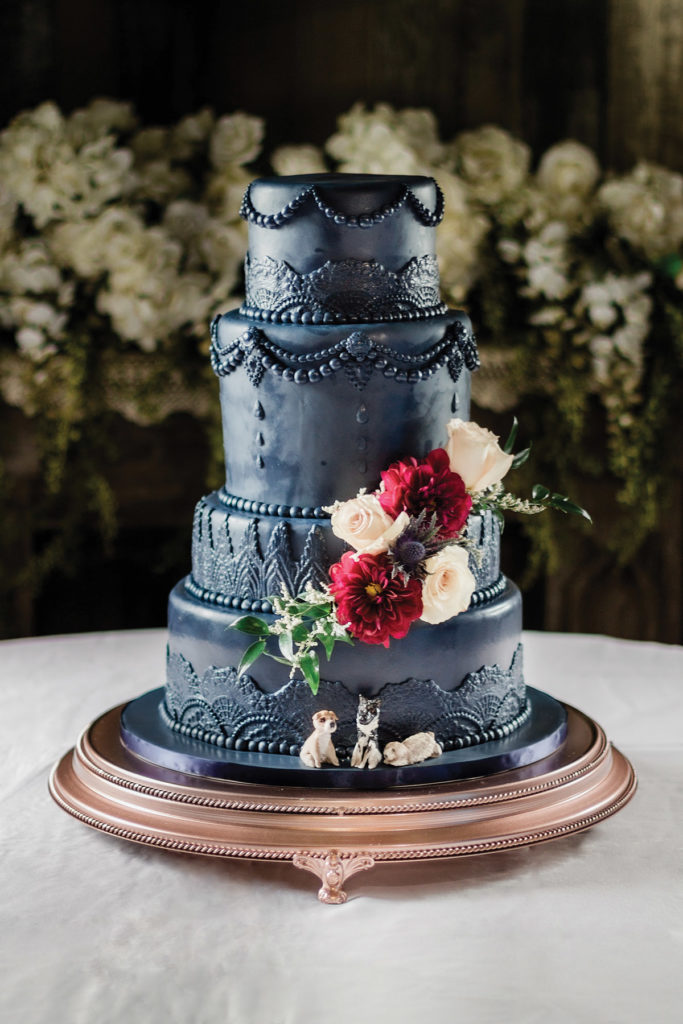 Four-tier navy blue cake with lace details, red and white flowers, and two dog figurines; blurred white flowers behind.