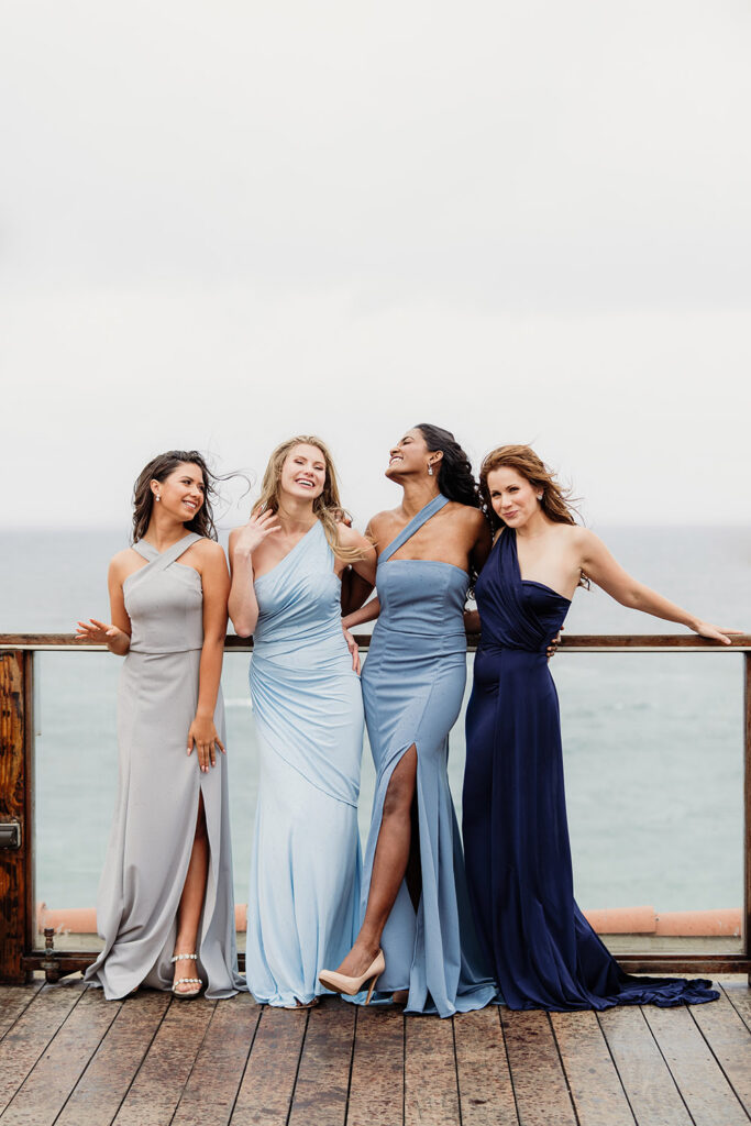 Four women in blue, gray, and navy gowns smile and pose on a wooden deck by the ocean under an overcast sky.