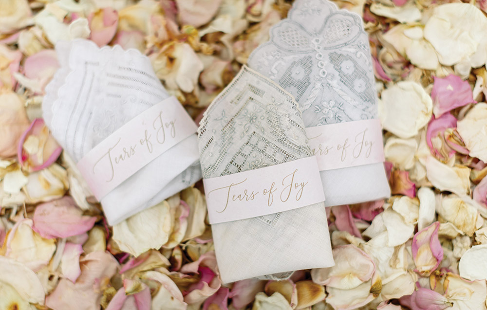 Three embroidered handkerchiefs labeled Tears of Joy sit on dried rose petals, featuring visible lace details.
