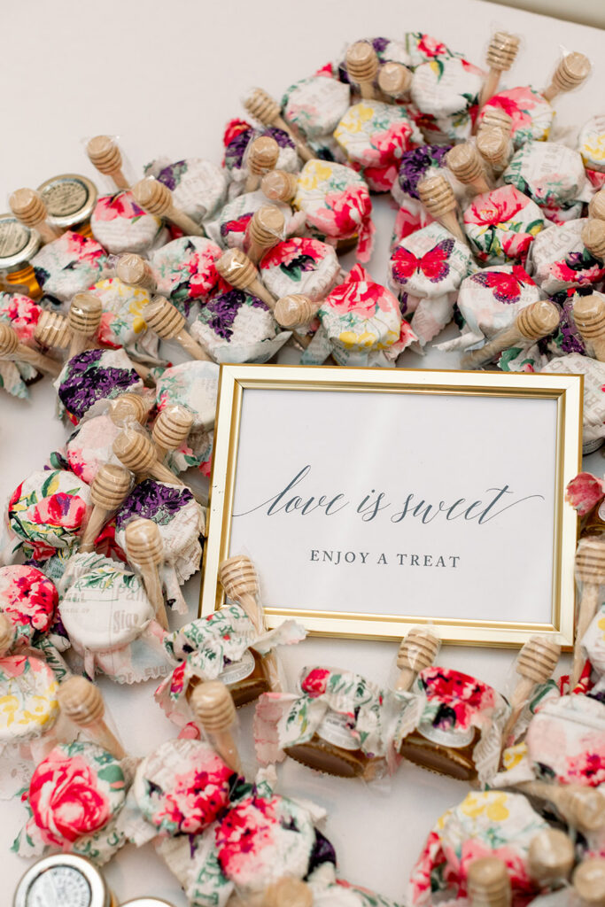 Honey jars with floral lids and dippers circle a framed “love is sweet, enjoy a treat” sign on a white surface.