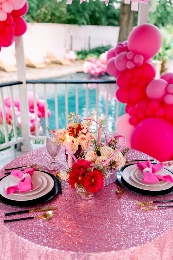 Round table with pink sequins, napkins, floral centerpiece, and elegant tableware by a pool with pink balloon arches.