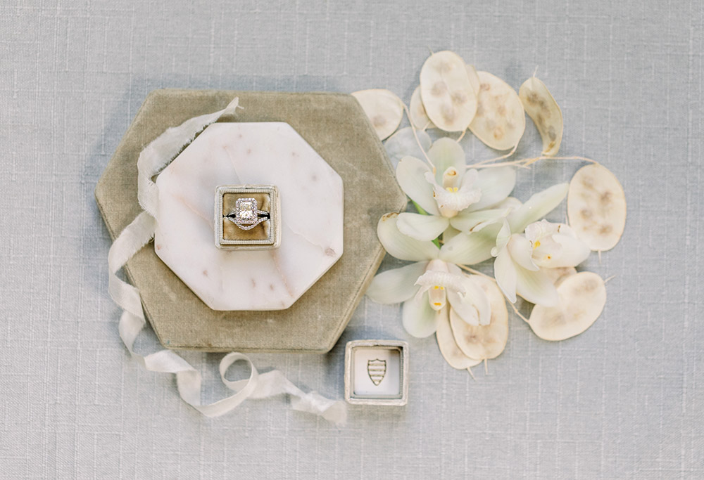 Engagement ring in hexagonal marble box, silver ribbon, white flowers, and shield ring box on light gray surface.