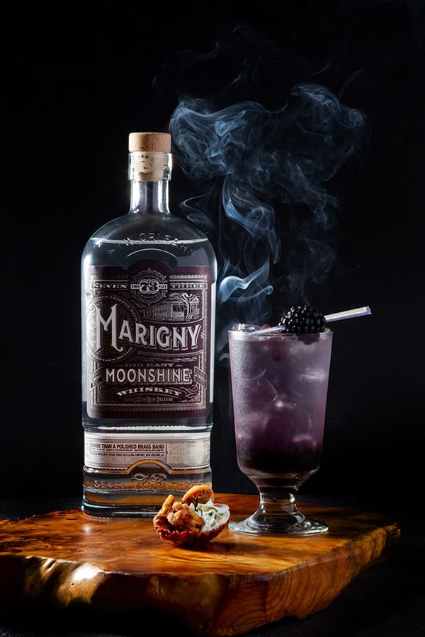 Marigny Moonshine bottle beside a smoky purple blackberry cocktail and pastry on wood, set against a dark background.