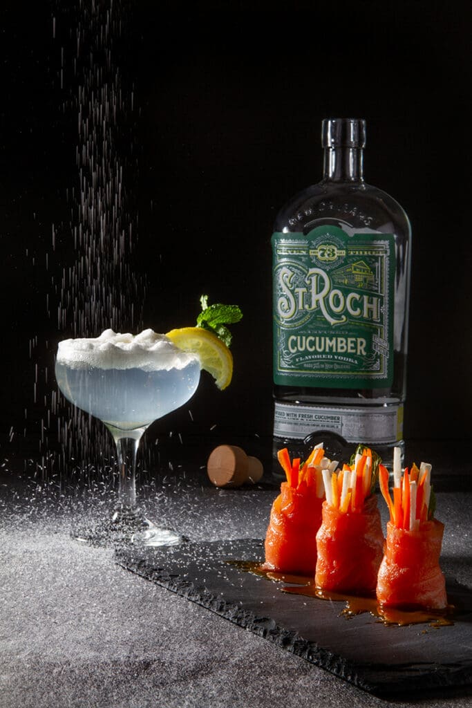 Foamy cocktail with lemon and mint next to St. Roch Cucumber gin, beside salmon veggie rolls on slate with salt or sugar sprinkled nearby.