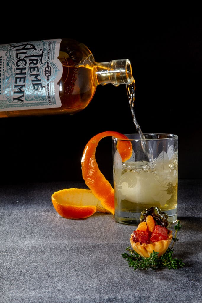 Bottle pouring liquor into iced glass with orange peel; small herb appetizer beside, on a dark background.
