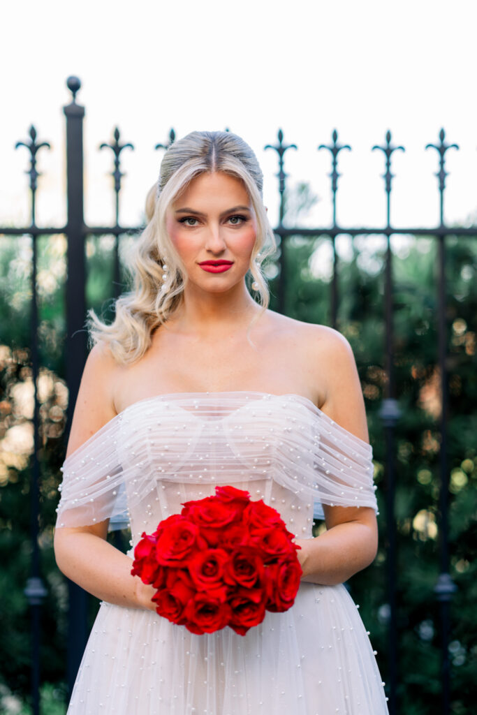 Bride in white off-shoulder dress with red roses, posing by a black iron fence and greenery, looking at the camera.