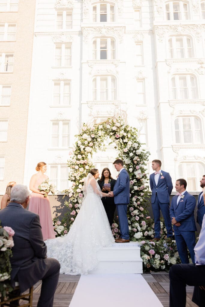 Bride and groom hold hands under a floral arch at their outdoor Louisiana wedding, with formal party and ornate building behind.