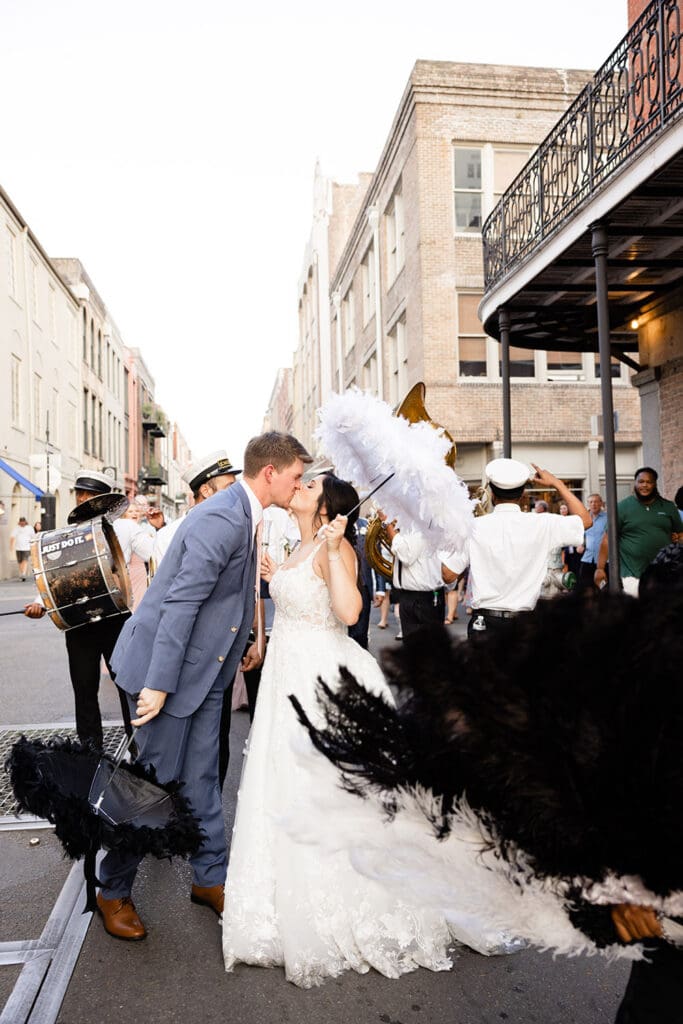 Bride and groom kiss in a New Orleans wedding parade, surrounded by musicians and fans with black and white feathers.