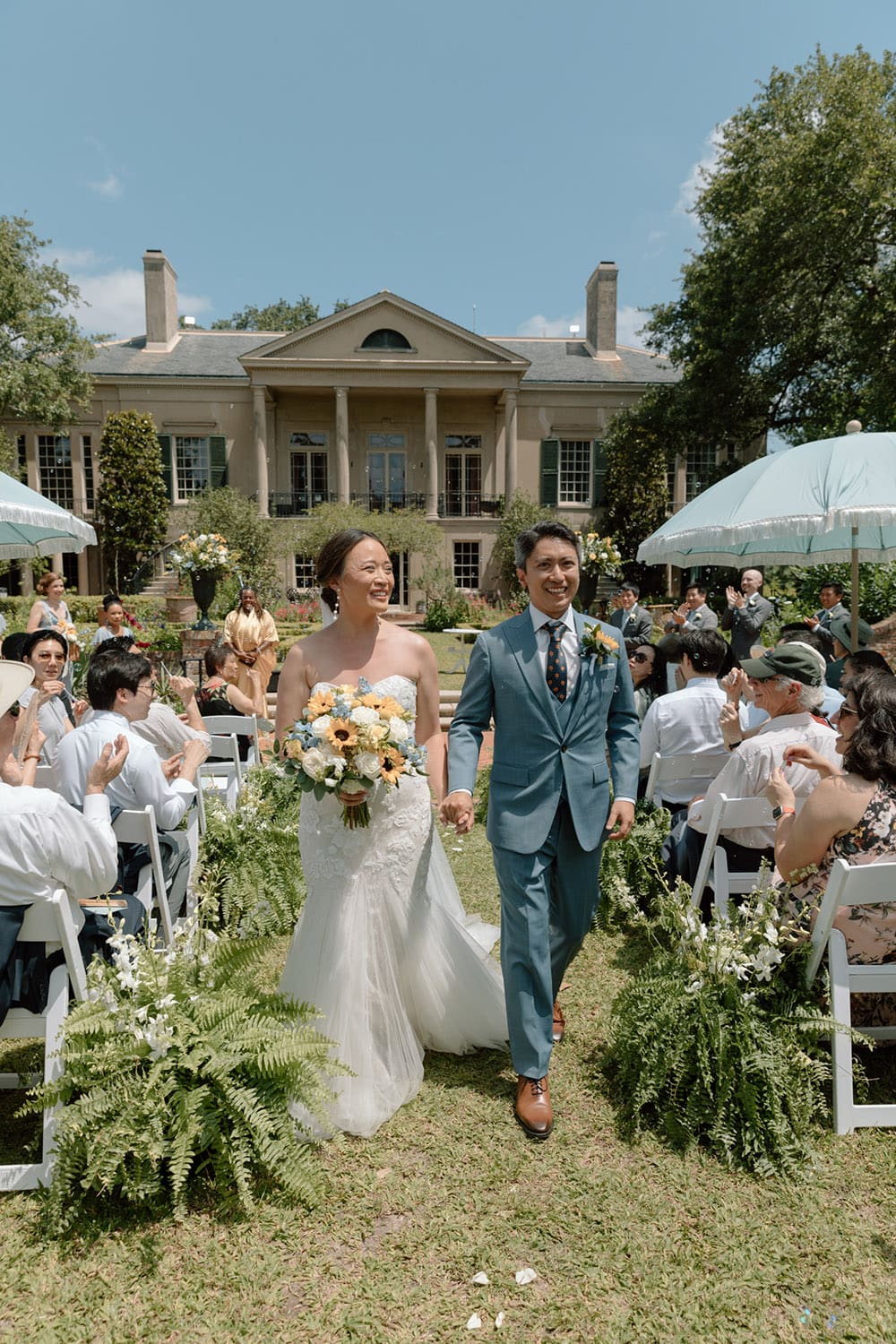 Smiling bride and groom walk hand in hand down the aisle at an outdoor Gulf South wedding by a large house.