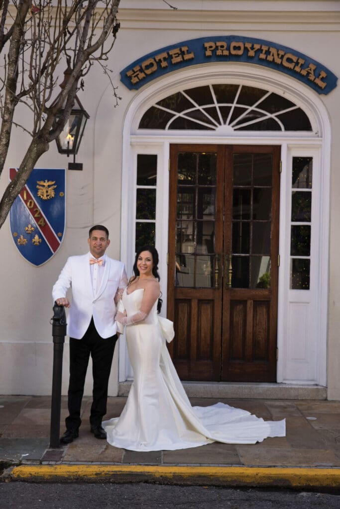 Bride and groom in formal attire pose by Hotel Provincial’s entrance, shield emblem, and double wooden doors in New Orleans.