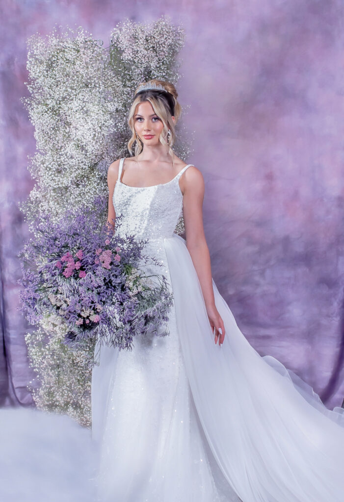 Bride in white gown holding purple and white bouquet before a pink and lavender backdrop with baby’s breath flowers.