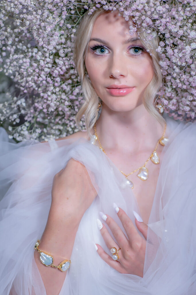 Blonde woman surrounded by white flowers, wearing a gold and pearl jewelry set and sheer white fabric.