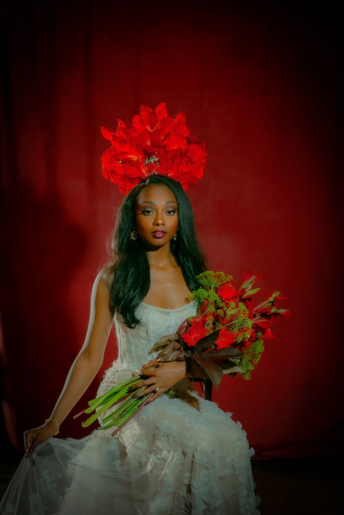 Woman in white dress with red floral headpiece holds red bouquet, seated before a red curtain.
