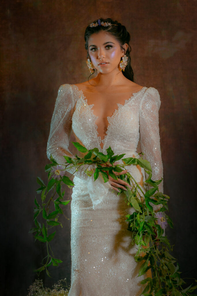 Woman in sparkling white gown with statement earrings holds floral arrangement against dark, textured background.