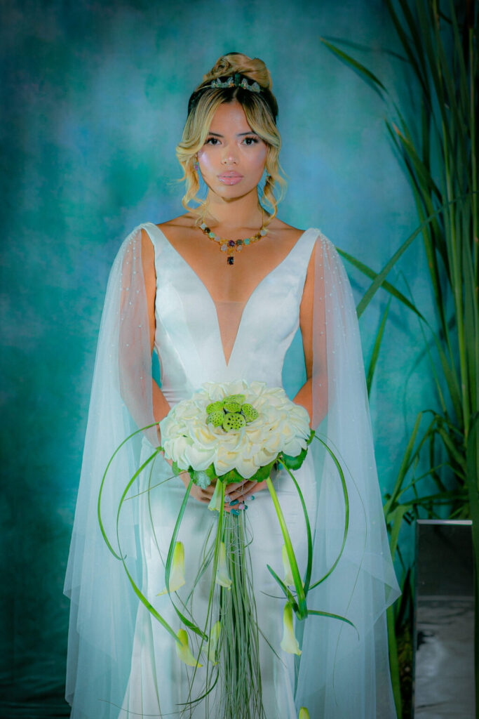 Woman in white dress with sheer cape sleeves holds white bouquet by a blue-green backdrop and tall plants.