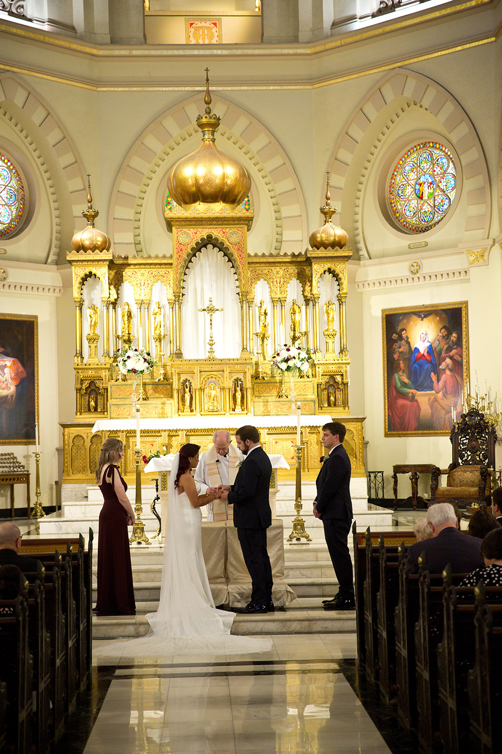 Bride and groom hold hands at a church altar during their wedding, with attendants nearby and gold-white decorations in the background.
