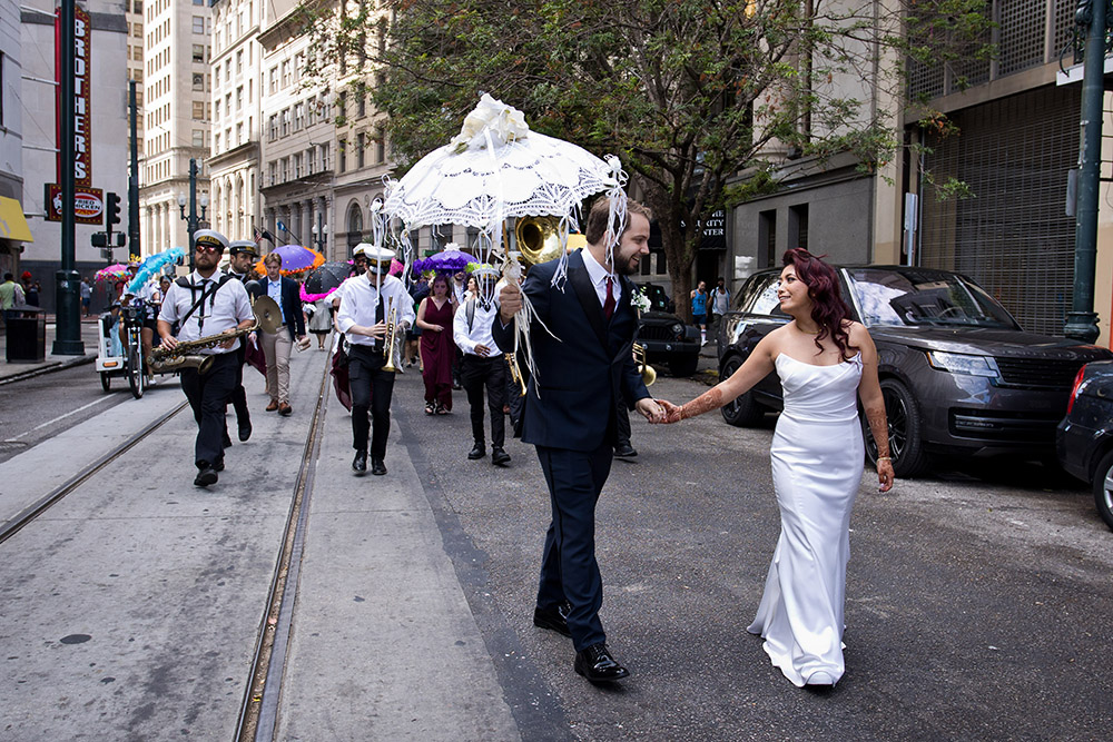 Bride and groom walk hand in hand down a city street, leading musicians and colorful umbrellas with a decorated white parasol.