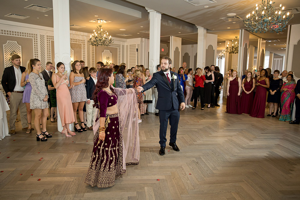 A couple in formal attire dances at a wedding as guests watch in a chandelier-lit, elegantly decorated venue with wood floors.