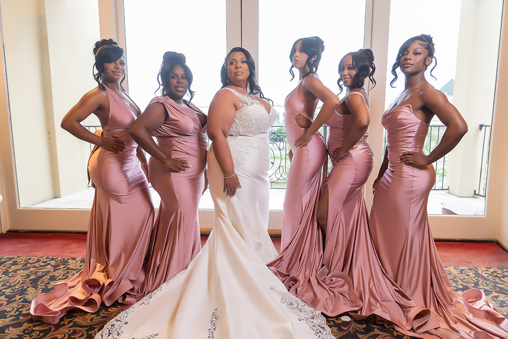 Bride in white gown poses with five smiling bridesmaids in pink dresses indoors by large windows.
