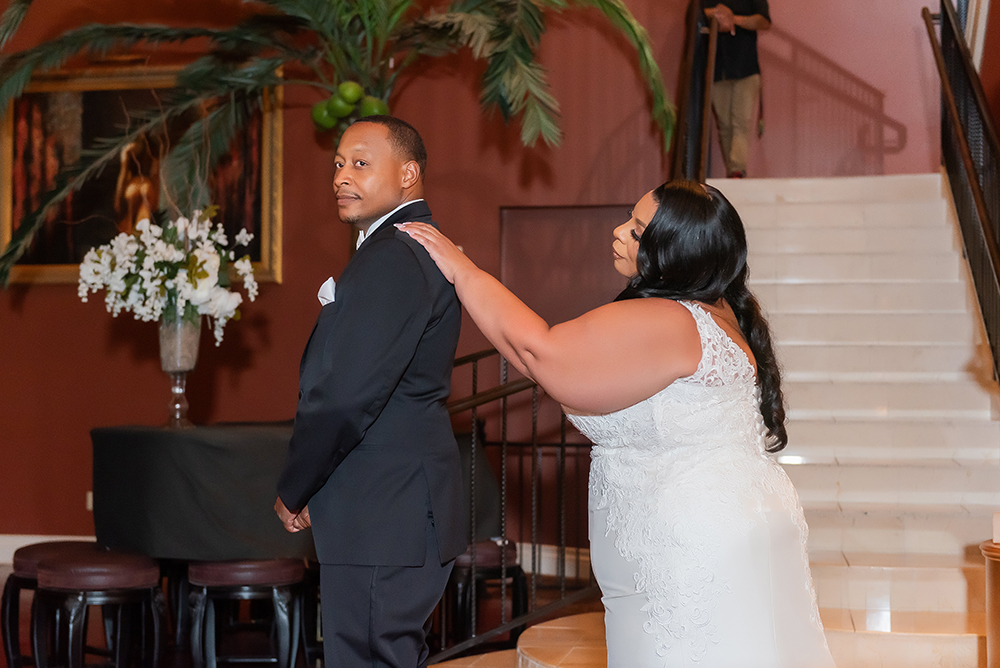 Bride in white lace gown touches groom’s shoulders from behind by a staircase with white flowers and a palm plant.