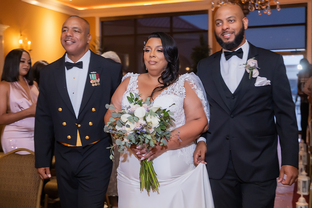 Bride in white with bouquet walks down aisle between two smiling men in formal wear; guests watch from seats and standing.