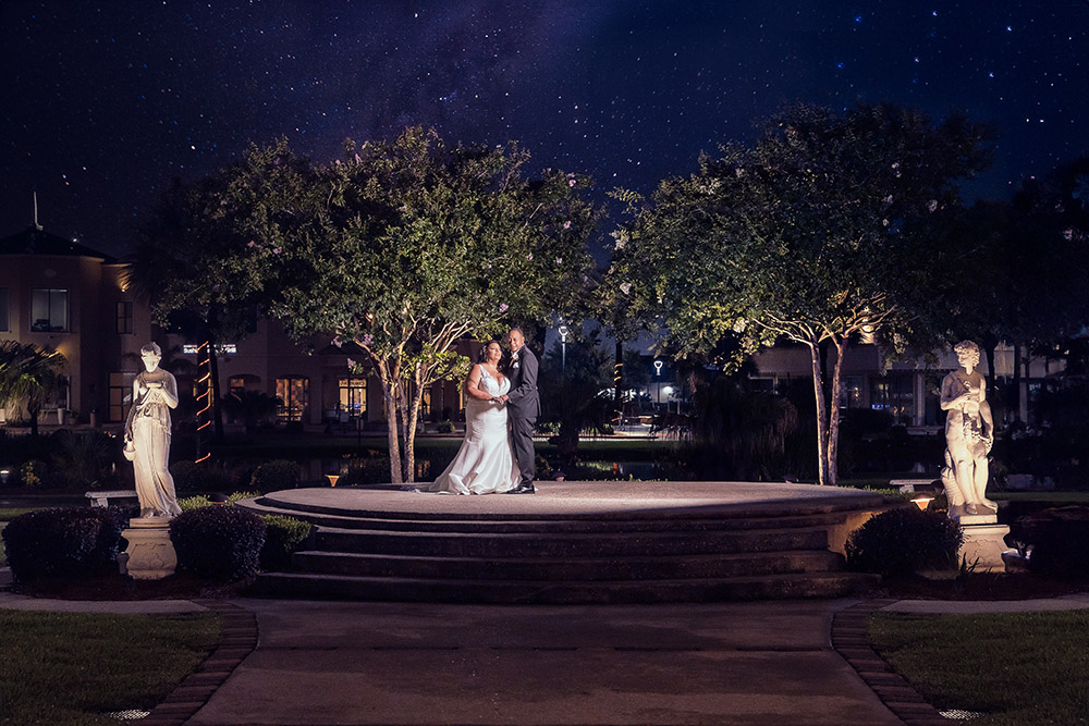 Bride and groom on an outdoor stone platform at night, with trees, statues, soft lighting, and a starry sky.