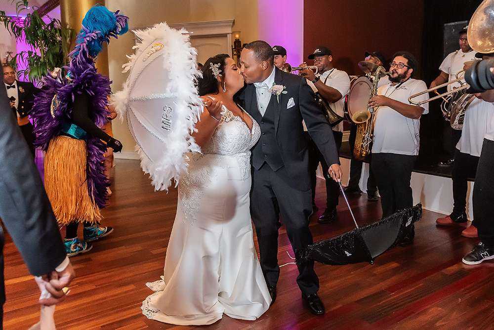 Bride and groom kiss under feathered umbrellas, leading a lively band and colorful dancers at an indoor wedding celebration.
