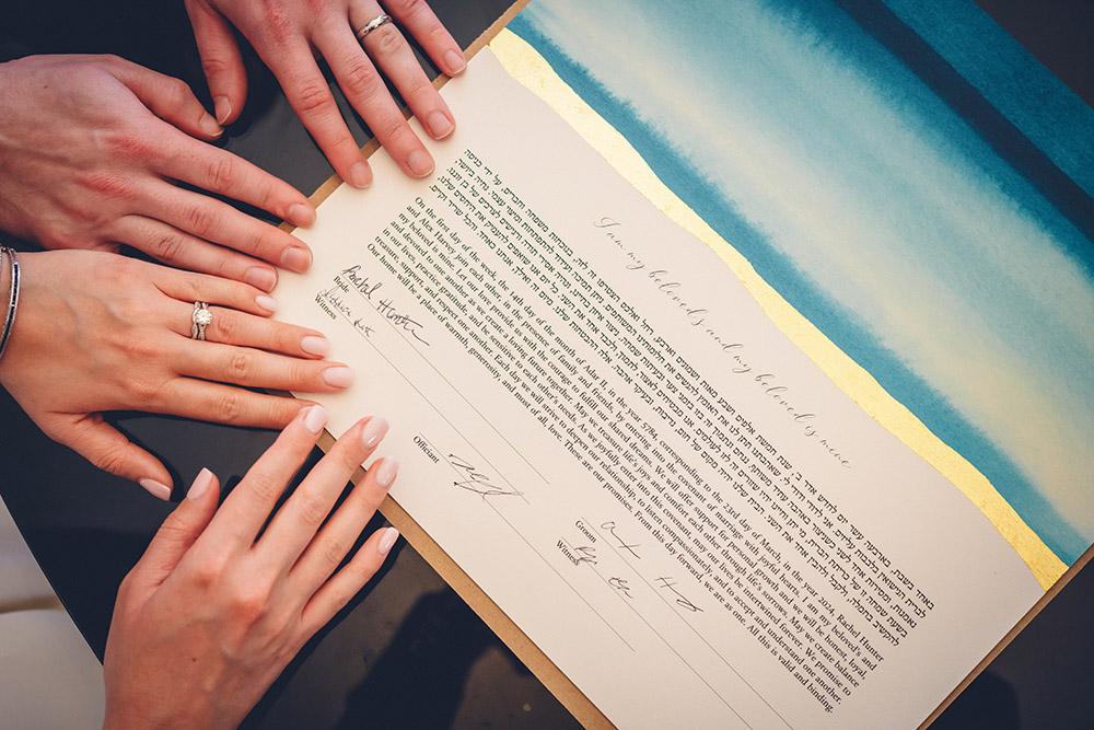 Three hands with wedding rings rest on a decorated ketubah with Hebrew text and English signatures at a wedding ceremony.