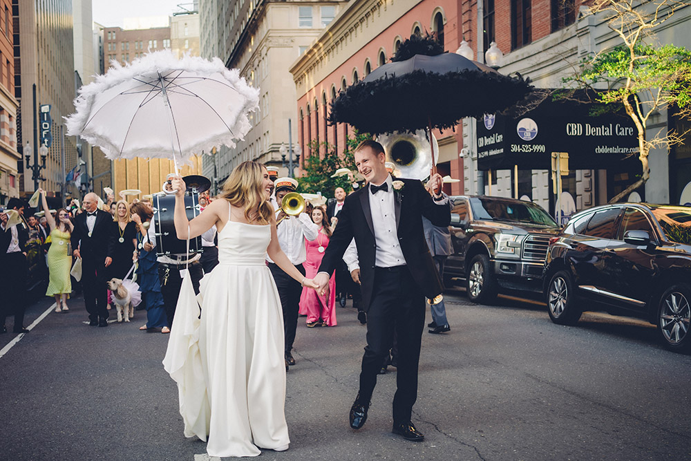 Bride and groom with decorated umbrellas lead a joyful wedding parade down a city street, followed by guests and musicians.