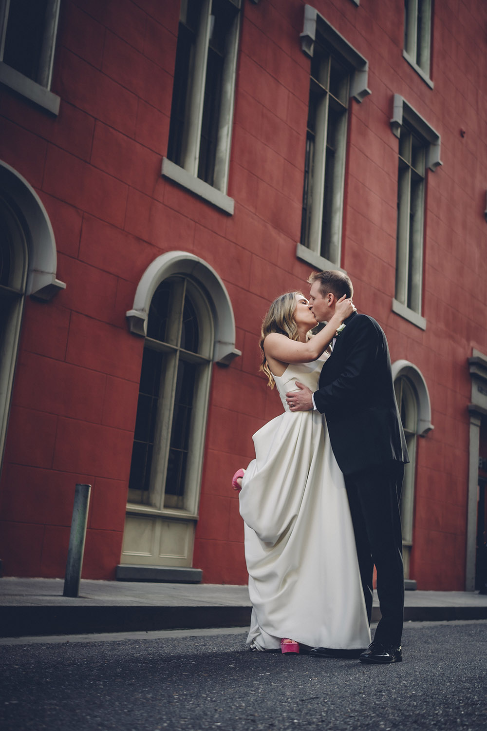 Bride in white dress and pink shoes kisses groom in black suit before a red building with tall windows.
