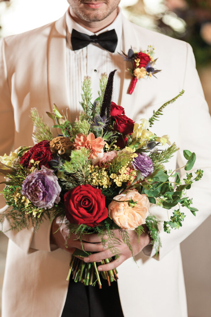 Man in a white tuxedo and black bow tie holding a bouquet of red, purple, and peach flowers with greenery.