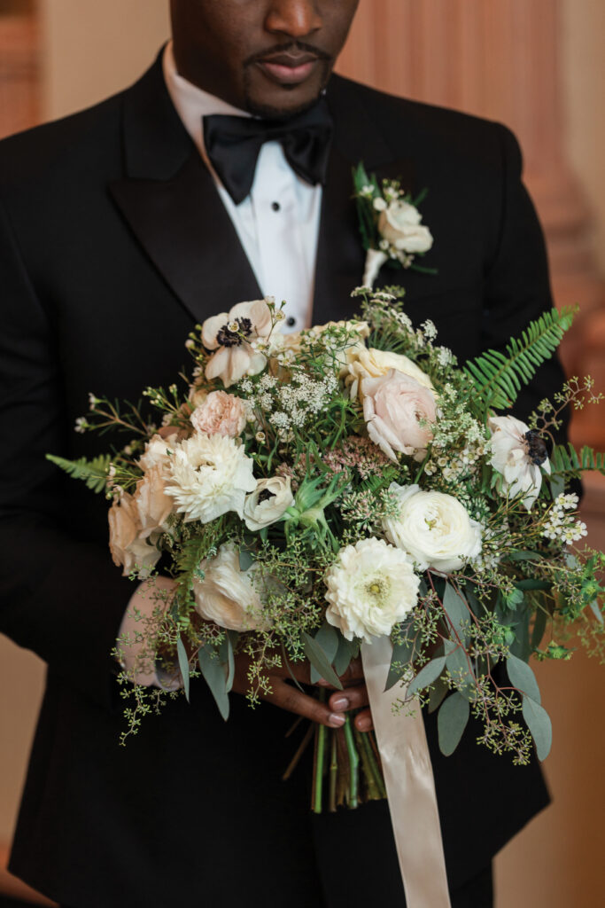 Man in black tuxedo holding a large bouquet of white and pale pink flowers with greenery and cream ribbon.