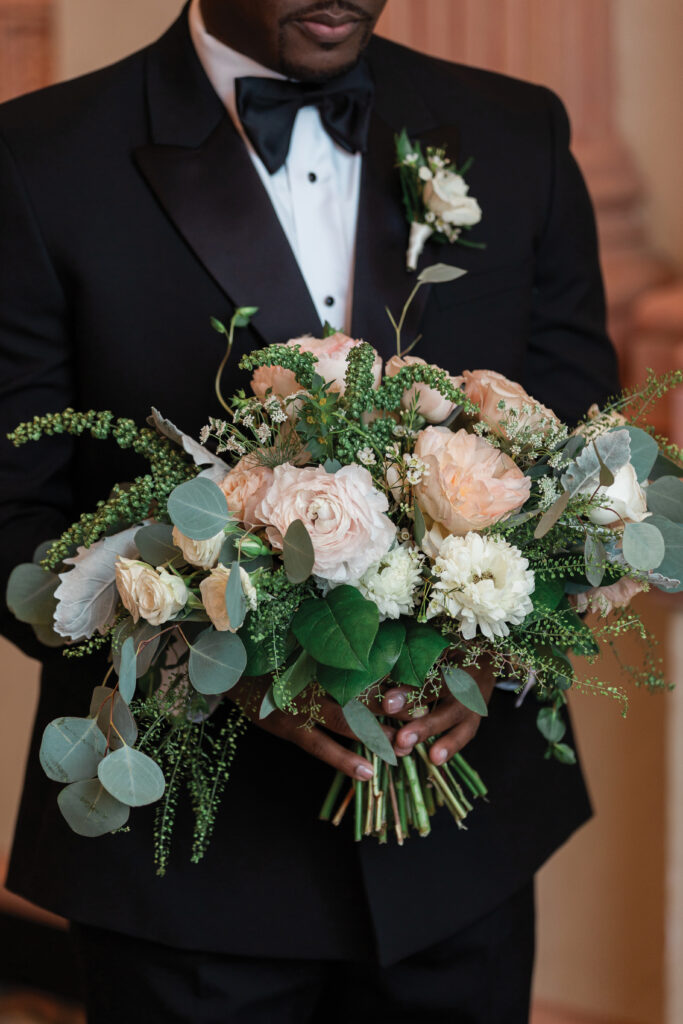 Person in a black tuxedo holding a bouquet of pale pink and white flowers with greenery.